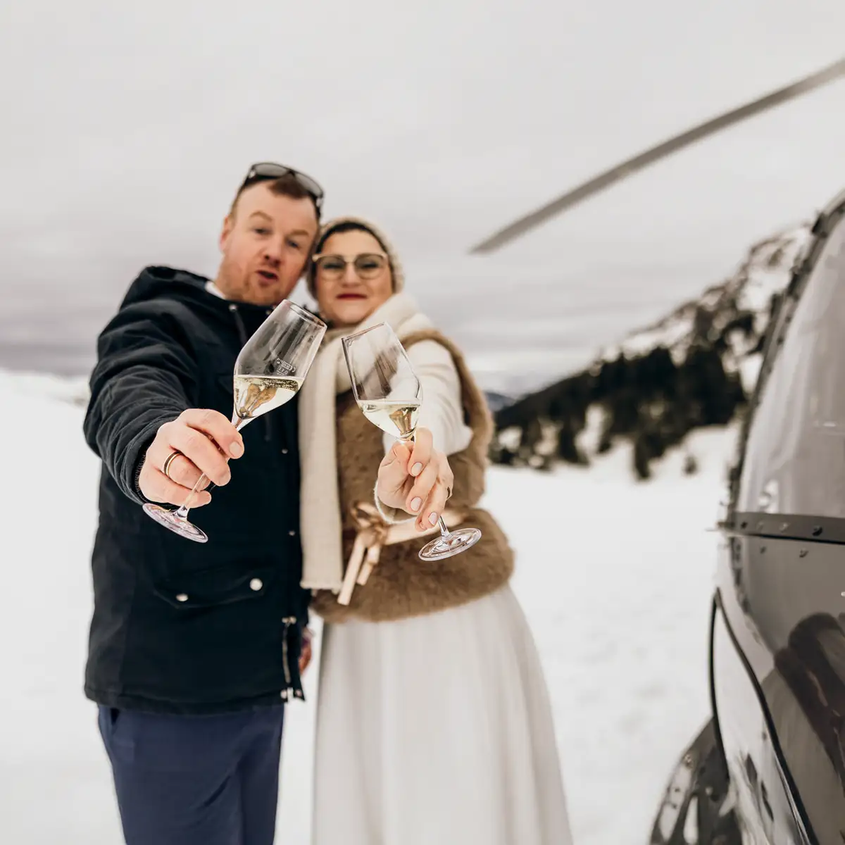 Ein frisch verheiratetes Paar stößt nach einem Hubschrauberflug mit einem Glas Champagner auf seine Trauung an. Die Winterhochzeit wurde von einer Hochzeitsplanerin organisiert und schafft eine intime, romantische Atmosphäre in verschneiter Landschaft.
