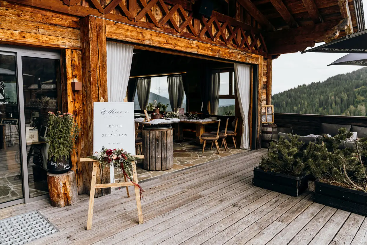 Willkommensschild zur Hochzeit auf der Alm vor rustikaler Berghütte mit Blick ins Tal und festlich gedecktem Innenbereich
