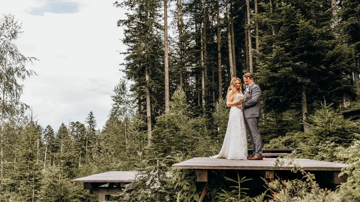Brautpaar steht auf einer Holzplattform inmitten eines dichten Waldes, umgeben von Tannen und natürlicher Idylle. Die Braut trägt ein elegantes Spitzenkleid, der Bräutigam einen grauen Anzug. Eine intime Hochzeitszeremonie in den Bergen unter freiem Himmel.